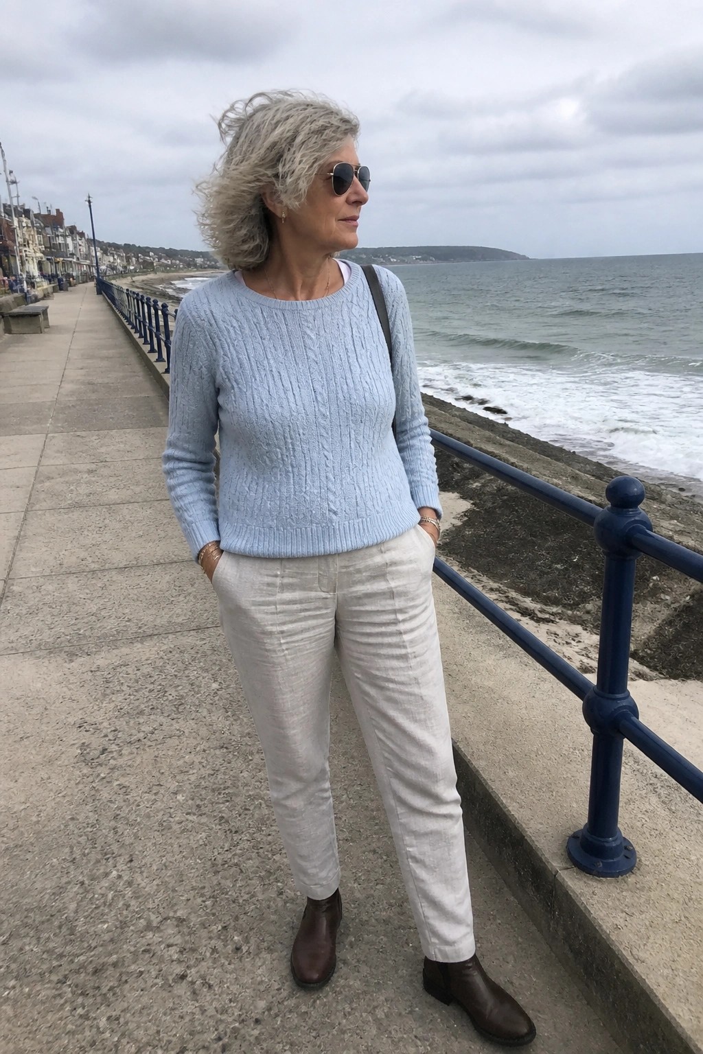 Woman with windswept gray hair in blue sweater and white pants by the sea