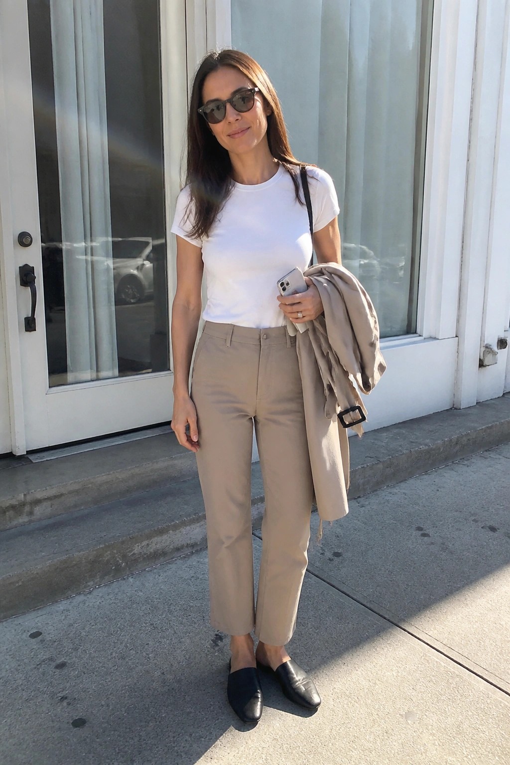 Woman in white t-shirt and beige pants holding phone and jacket outdoors