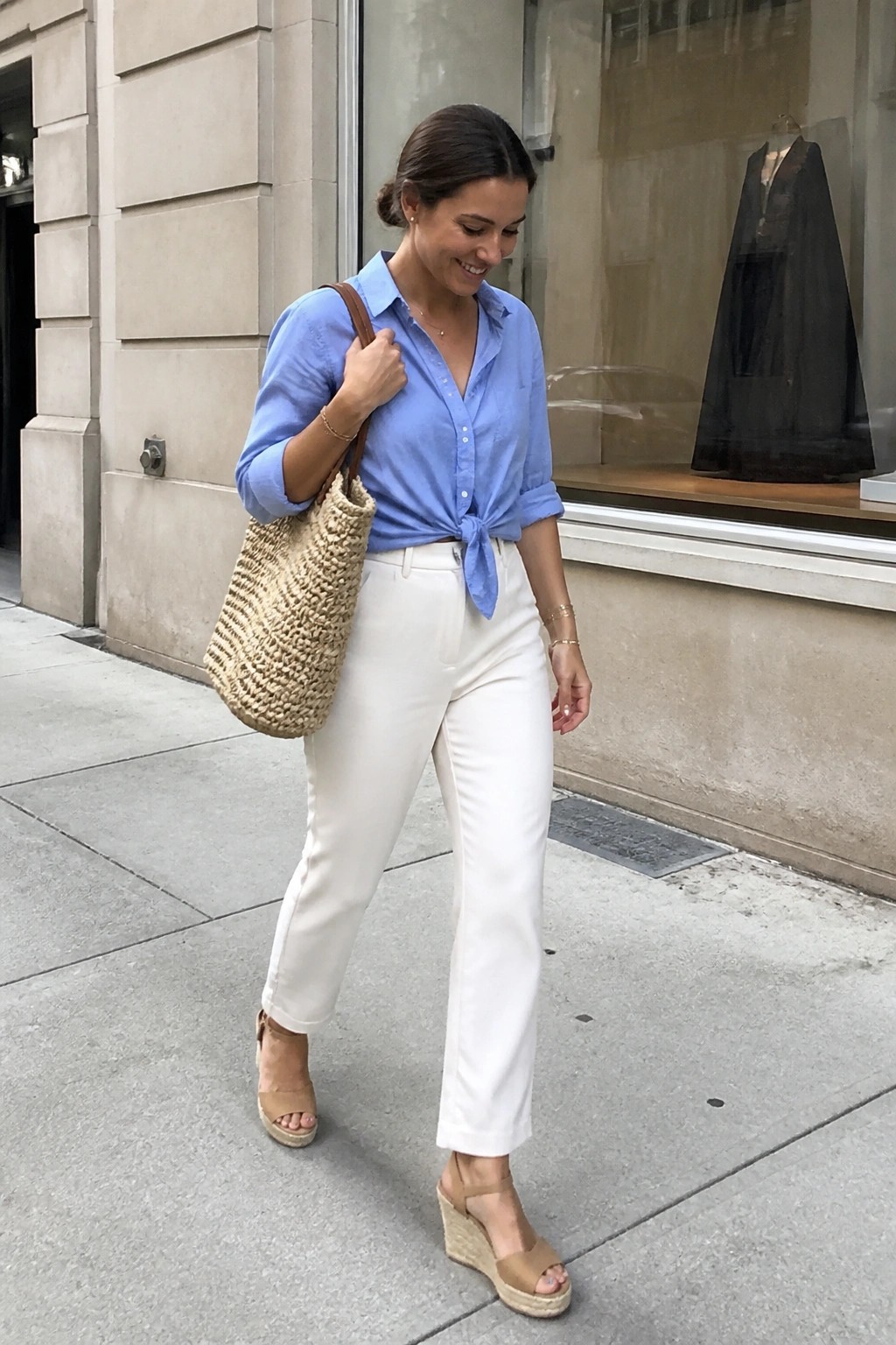 Woman in blue tied shirt and white pants carrying woven tote on sidewalk