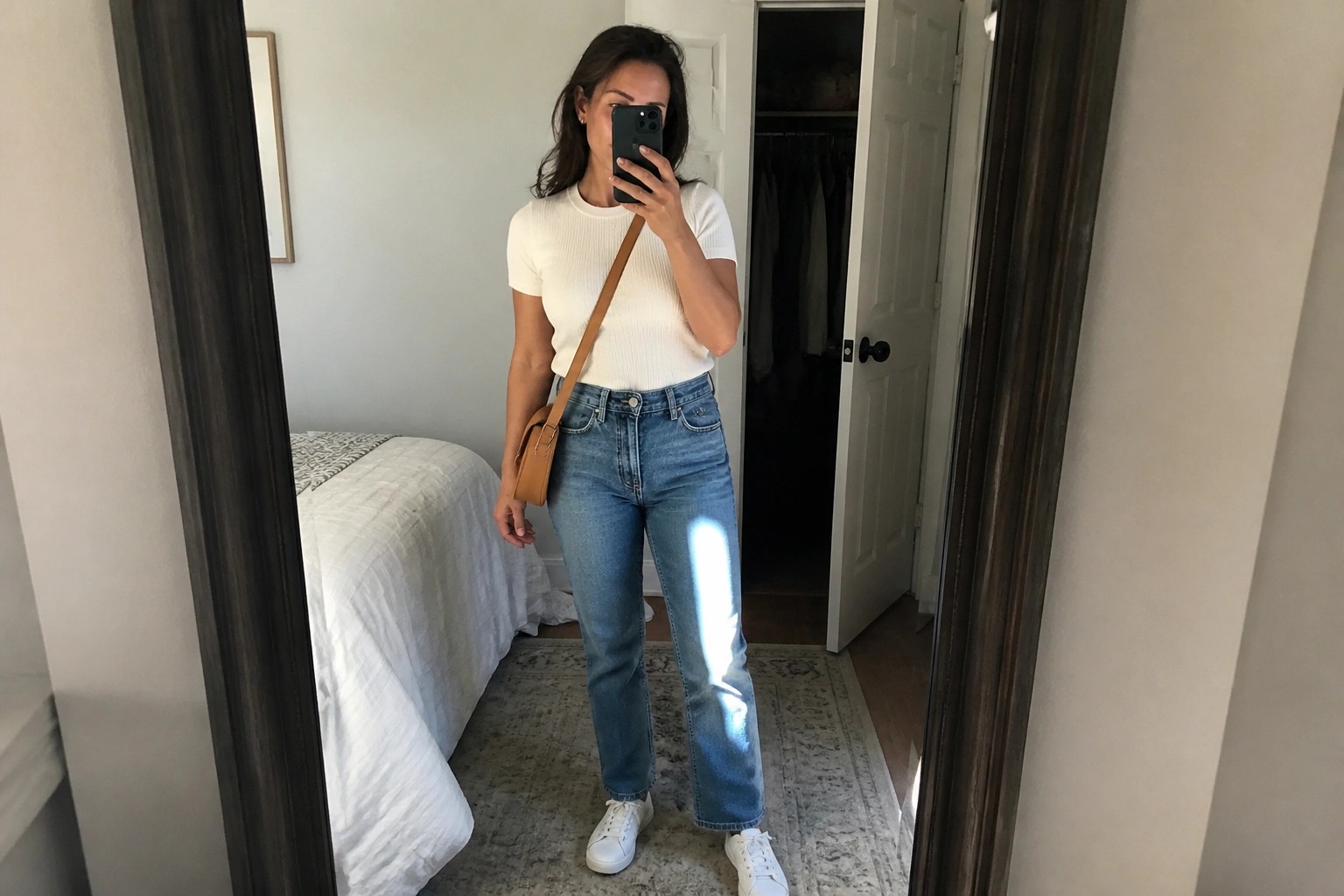 Woman in white top and blue jeans taking bedroom mirror selfie with phone