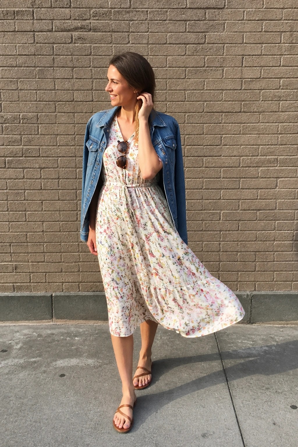 Woman in floral dress and denim jacket standing by brick wall