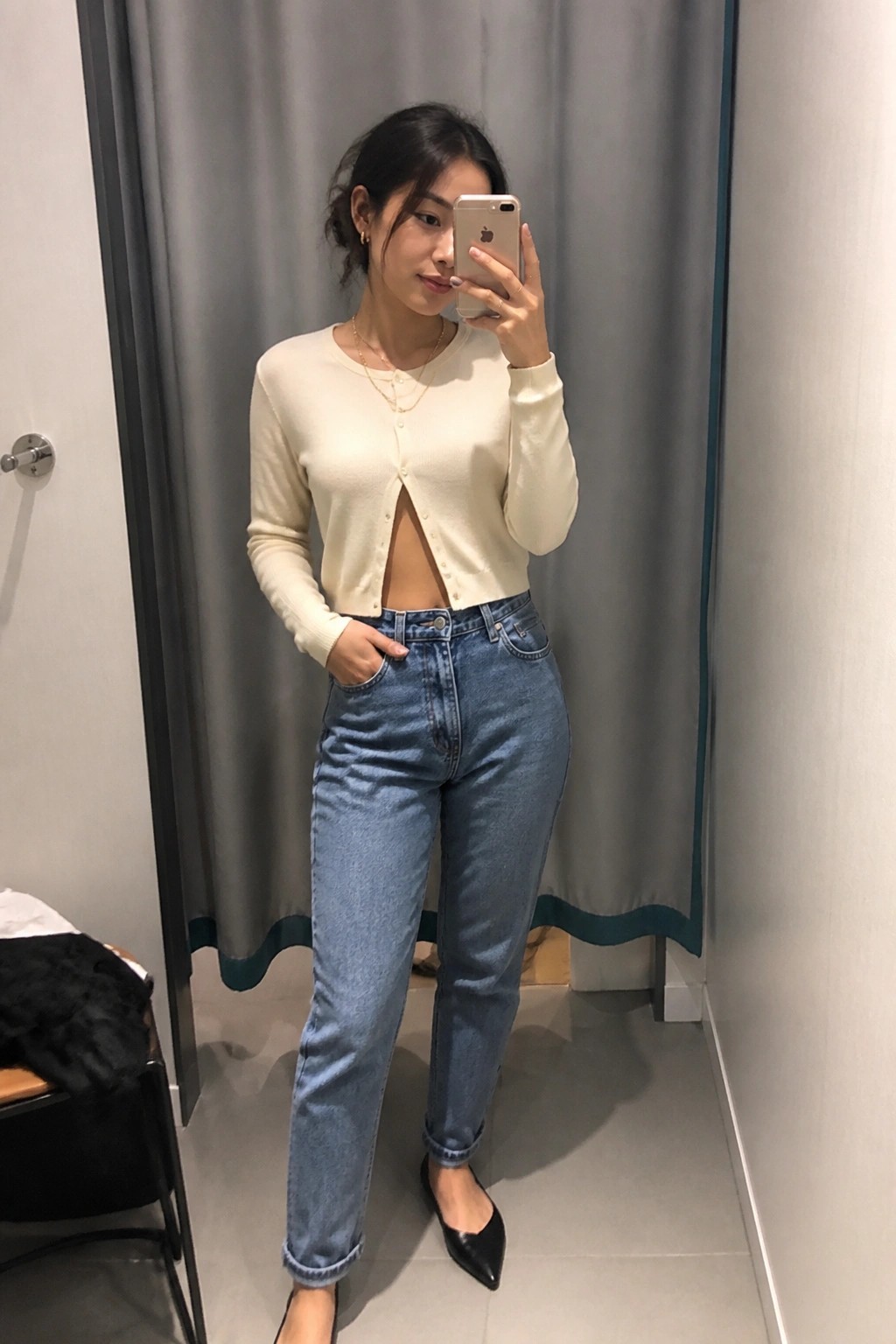 Woman wearing a cream cropped cardigan over high waisted blue jeans with black pointed flats and layered gold necklaces, captured in a mirror selfie.