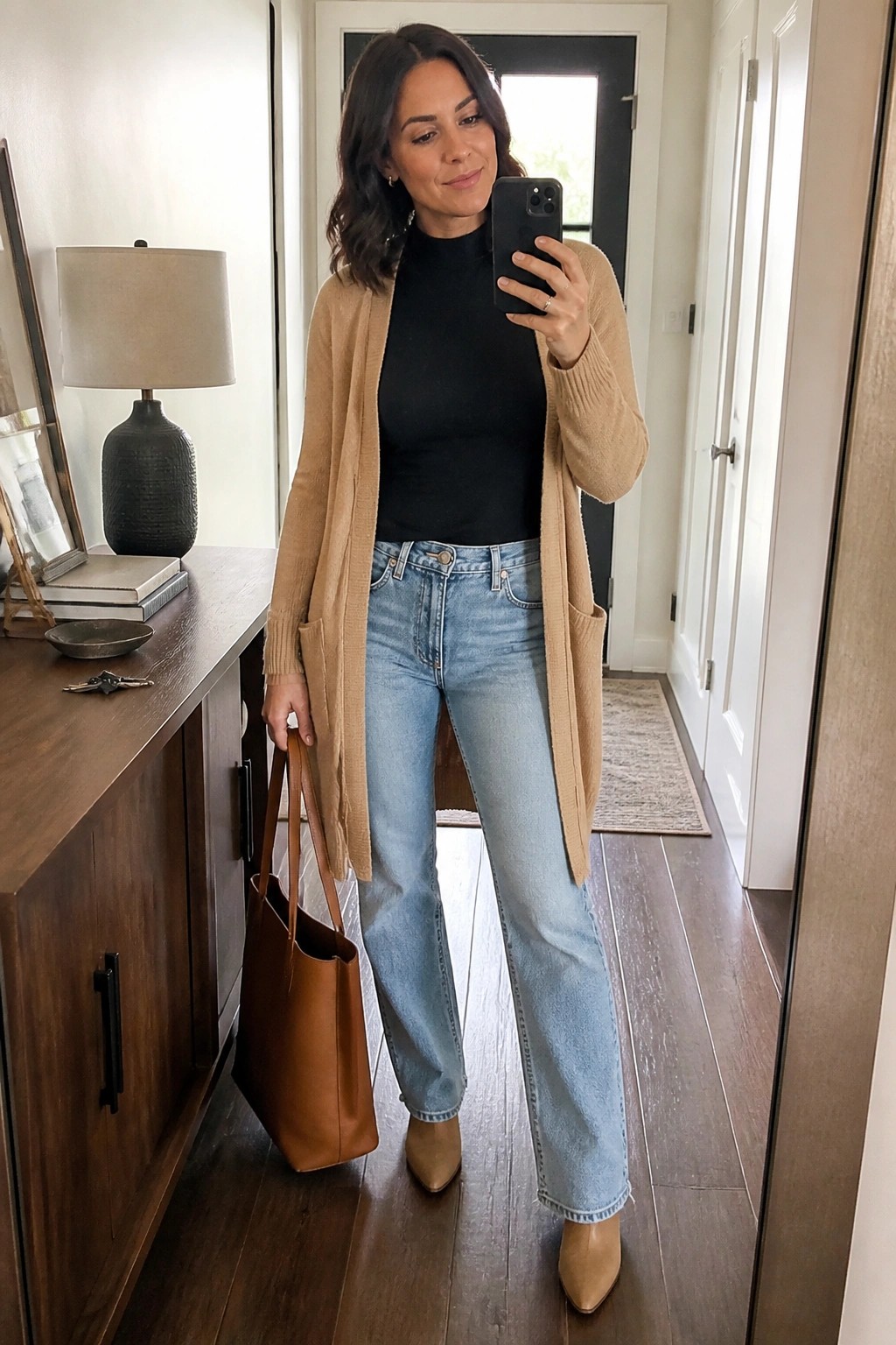 Woman in tan cardigan and jeans taking mirror selfie while holding brown tote bag