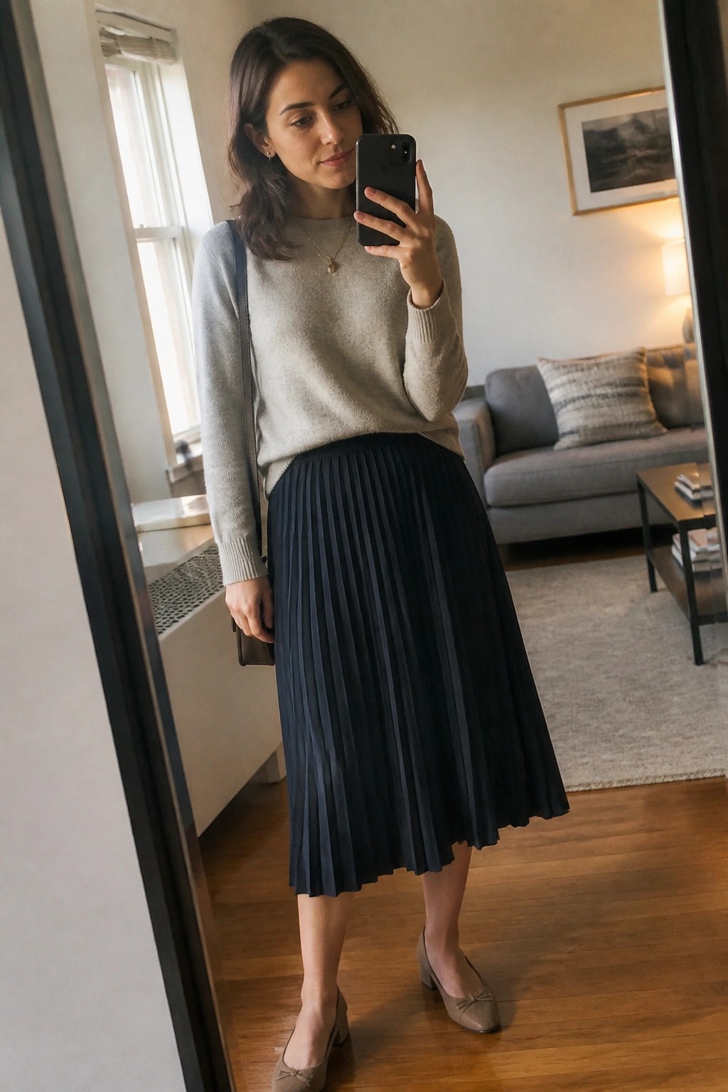 Woman in gray sweater and navy pleated skirt taking mirror selfie in living room