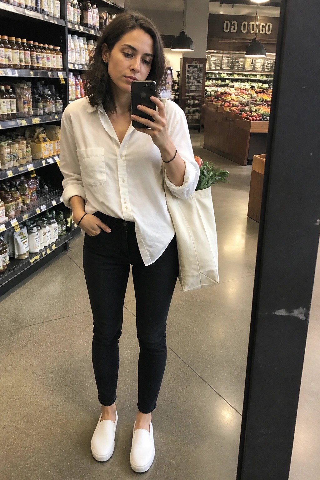 Woman wearing a white button-up shirt with black skinny jeans and white slip-on sneakers while holding a canvas tote bag.