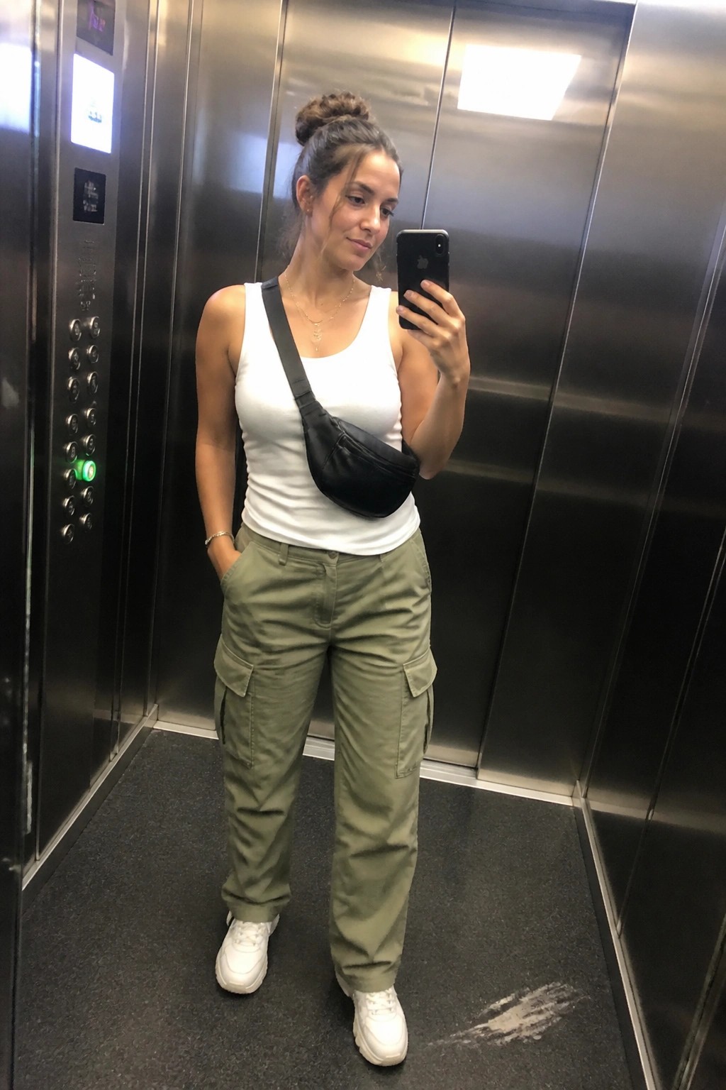 Woman in white tank top and green cargo pants taking selfie in elevator