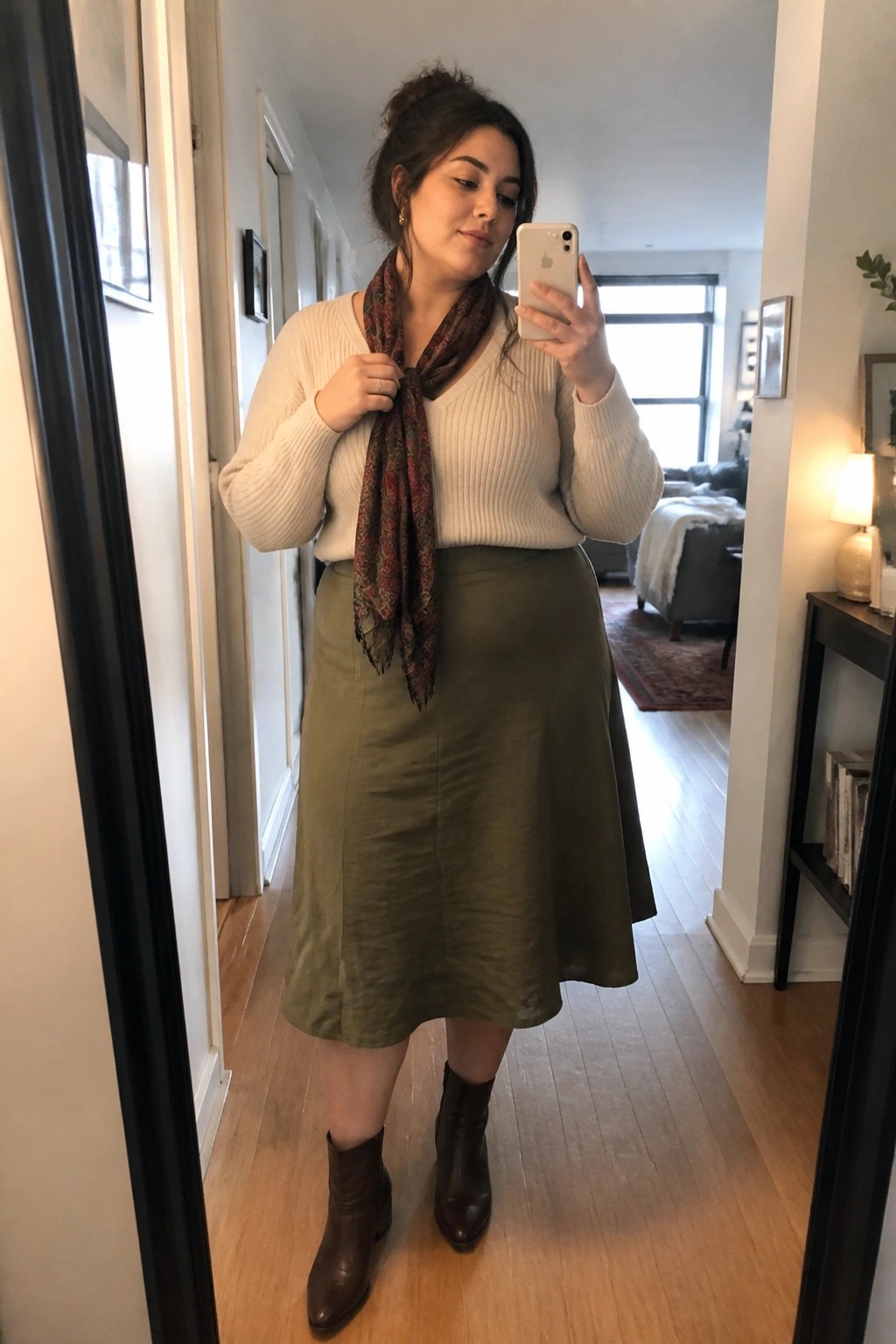 Woman taking mirror selfie in cream sweater, olive skirt, and scarf