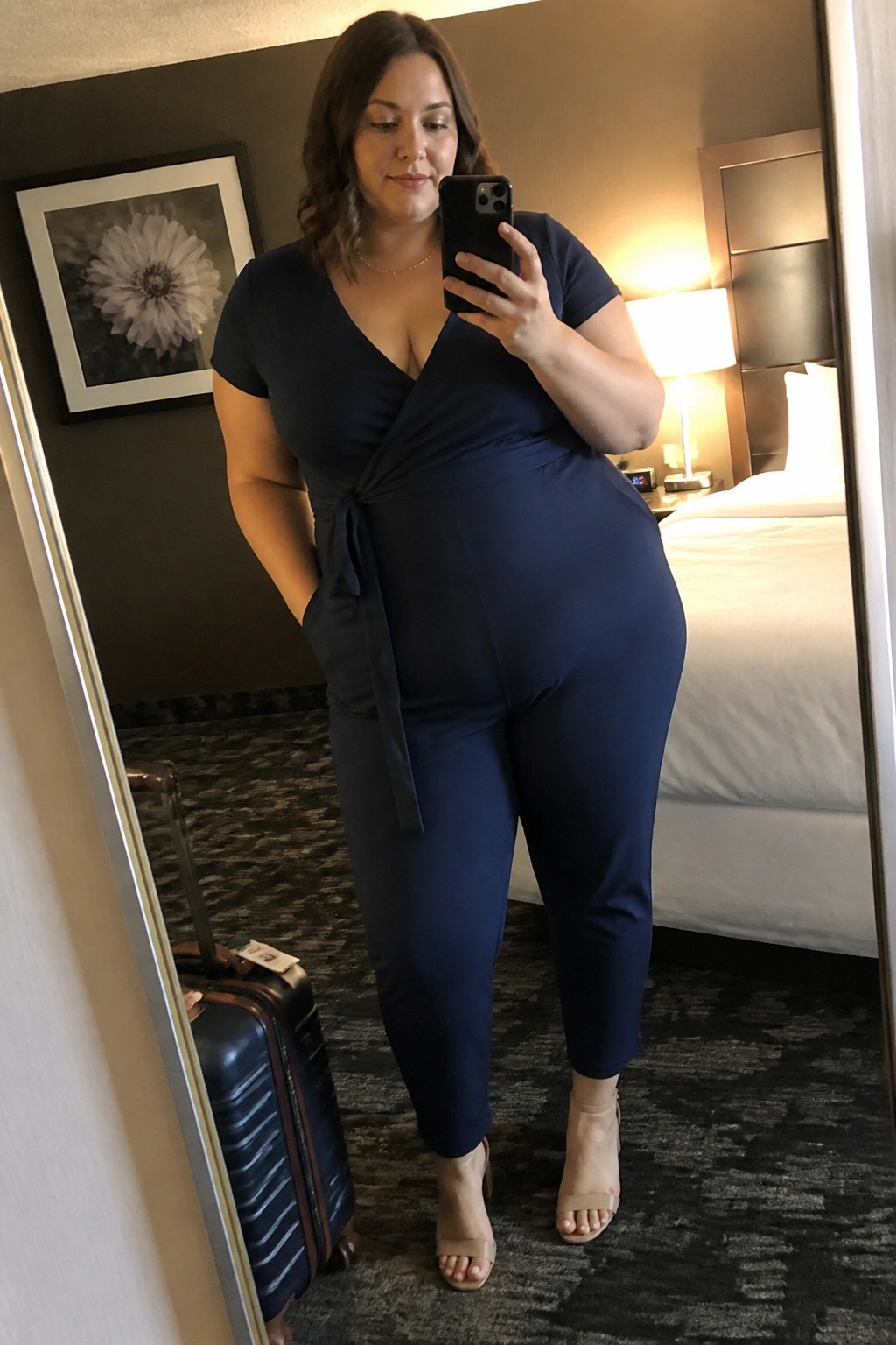 Woman in navy blue jumpsuit taking mirror selfie in hotel room with suitcase