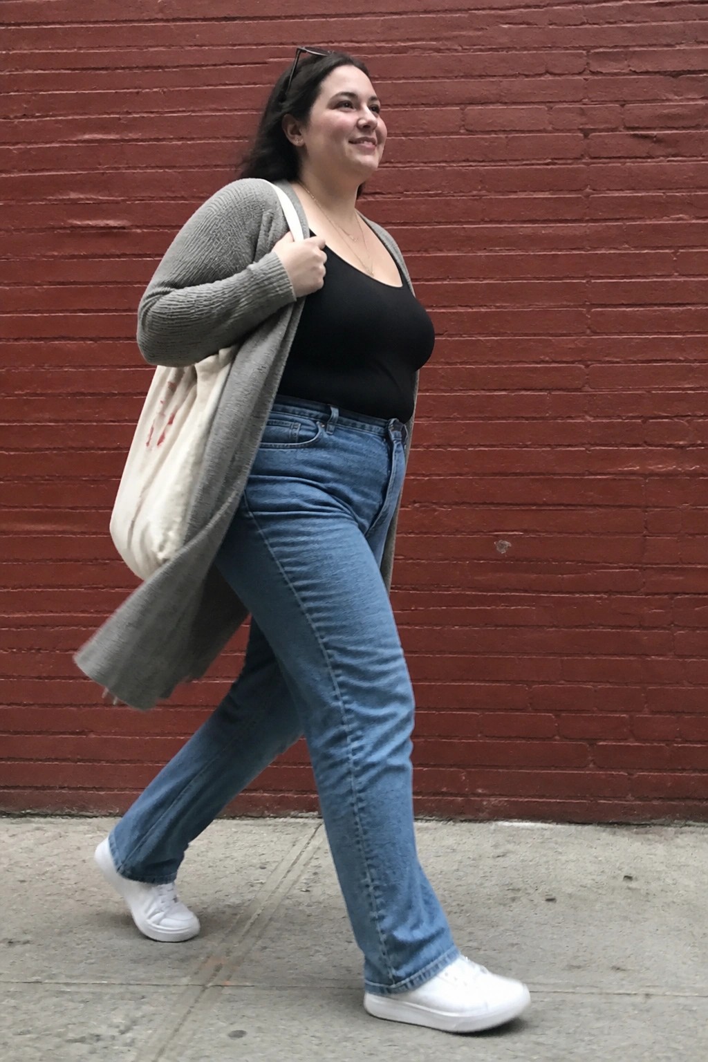 Woman in blue jeans and gray cardigan carries tote bag past red brick wall