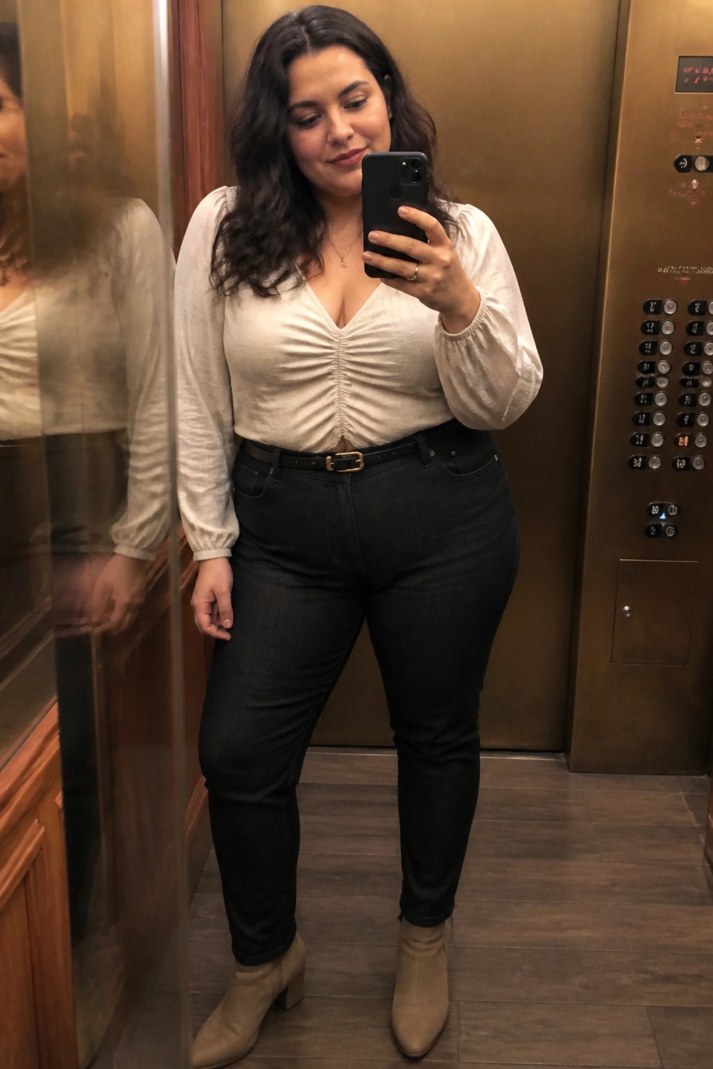 Woman in white ruched top and black jeans taking elevator mirror selfie
