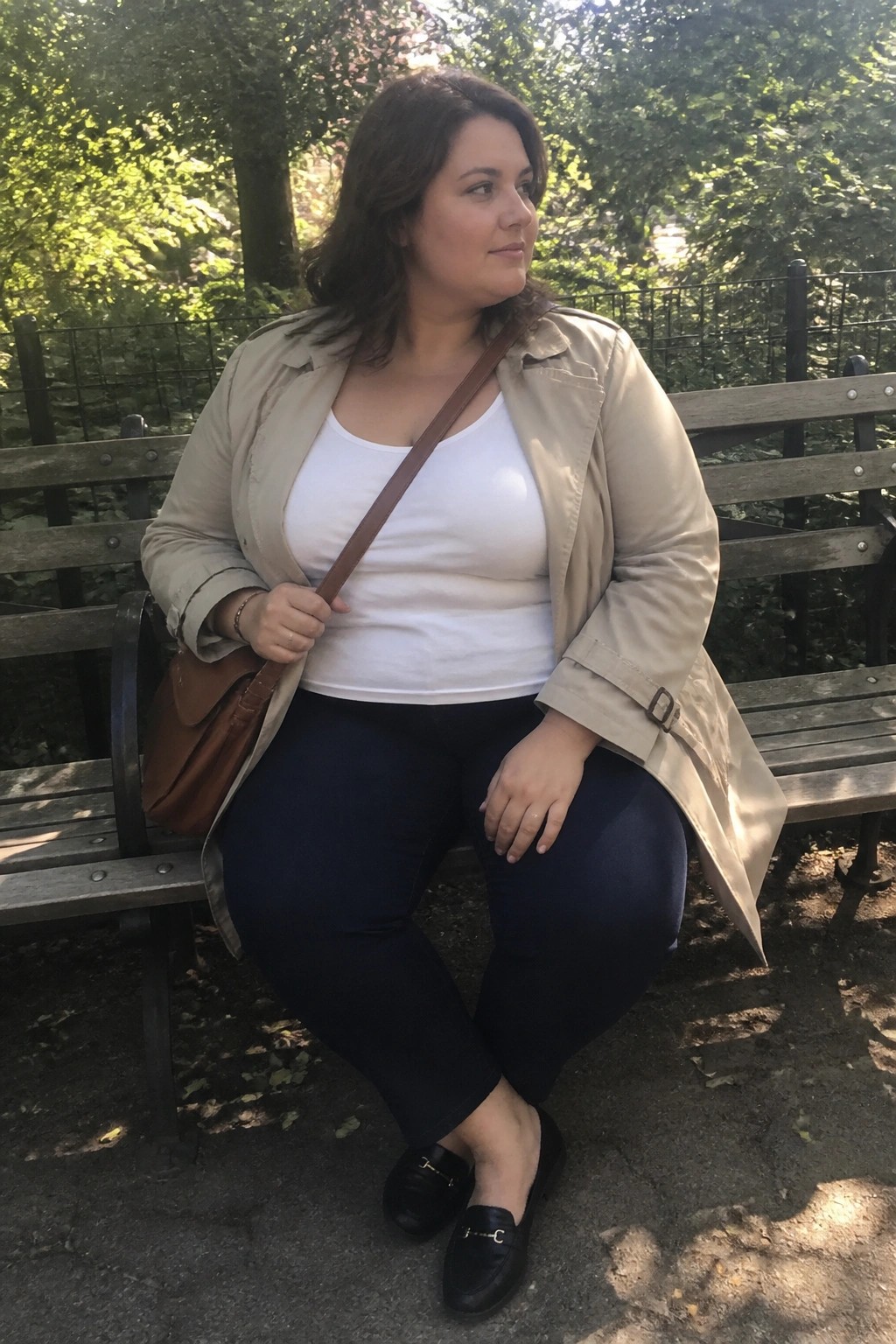 Plus size woman seated on bench wearing open beige trench coat over white tank top with dark navy pants black loafers and brown crossbody bag.
