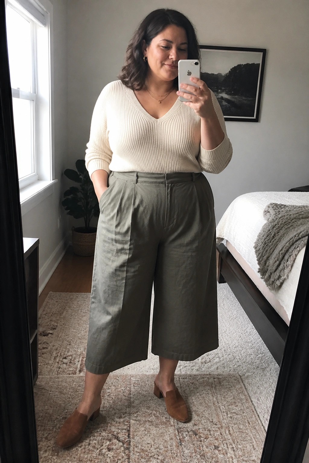 Plus size woman wearing olive green wide leg cropped pants with a cream ribbed V-neck sweater, brown suede mules, and layered gold necklaces while taking a mirror selfie.