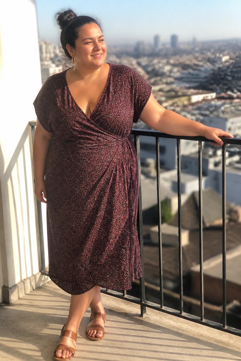 Smiling woman in sparkly burgundy wrap dress on sunny city balcony.