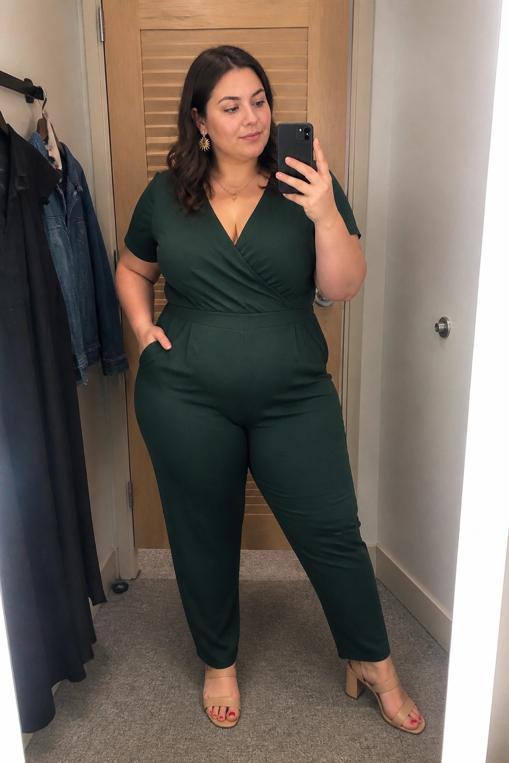 Woman in dark green jumpsuit taking mirror selfie in dressing room wearing beige heels