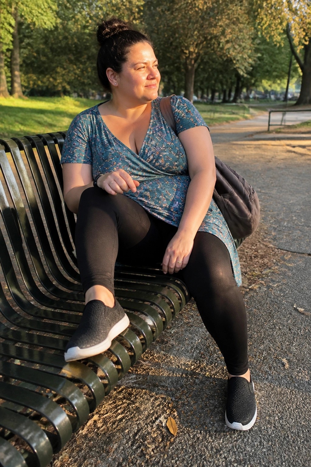 Plus size woman wearing a teal floral wrap top with short sleeves over black leggings and black slip on sneakers while seated on a park bench.