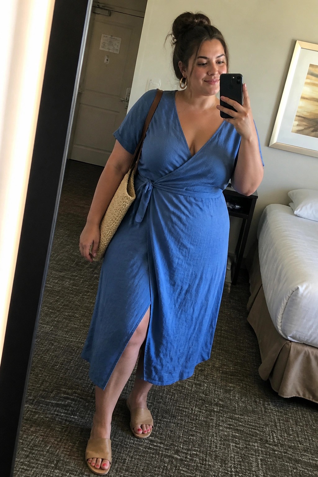Woman in blue wrap dress taking mirror selfie in hotel room with woven bag