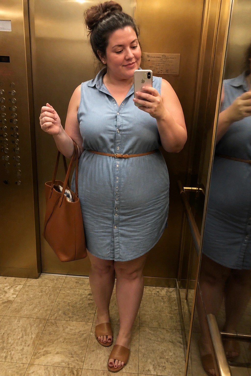 Woman in light blue sleeveless button-down denim shirt dress cinched with a narrow brown belt, carrying a brown leather tote and wearing matching slide sandals while taking a mirror selfie.