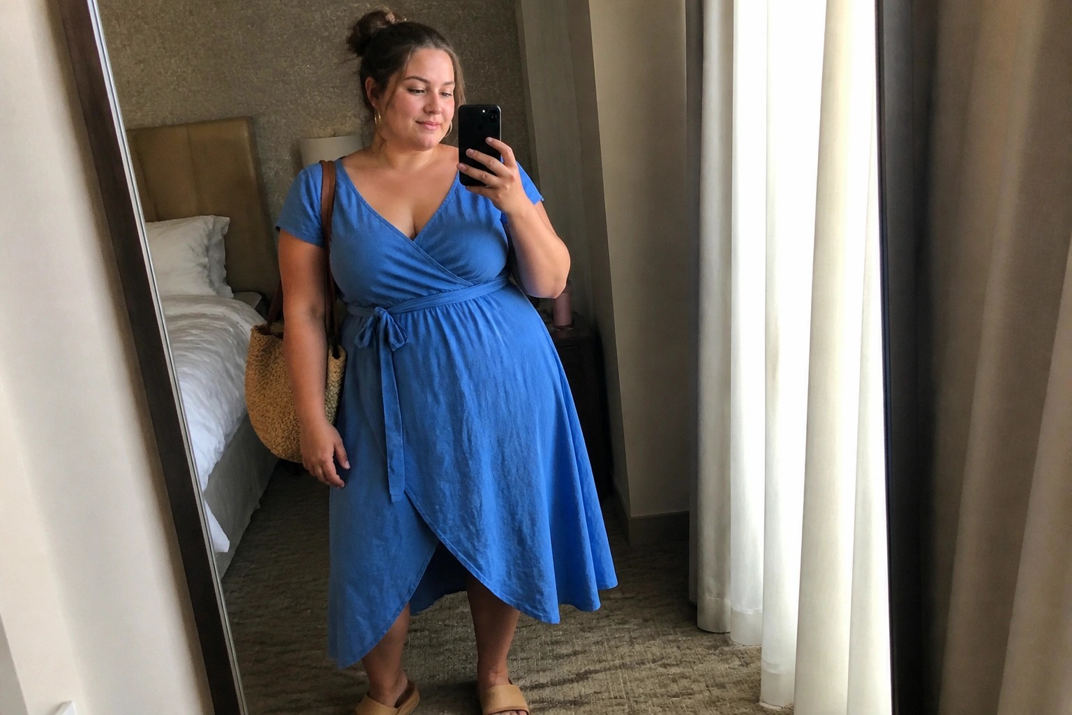 Woman in blue wrap dress taking mirror selfie in hotel room with woven bag