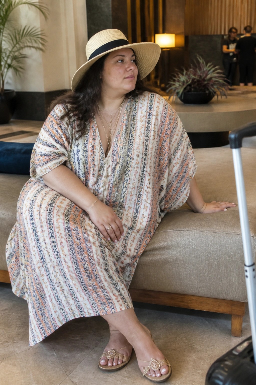 Woman in patterned caftan and straw hat seated on lobby couch beside suitcase