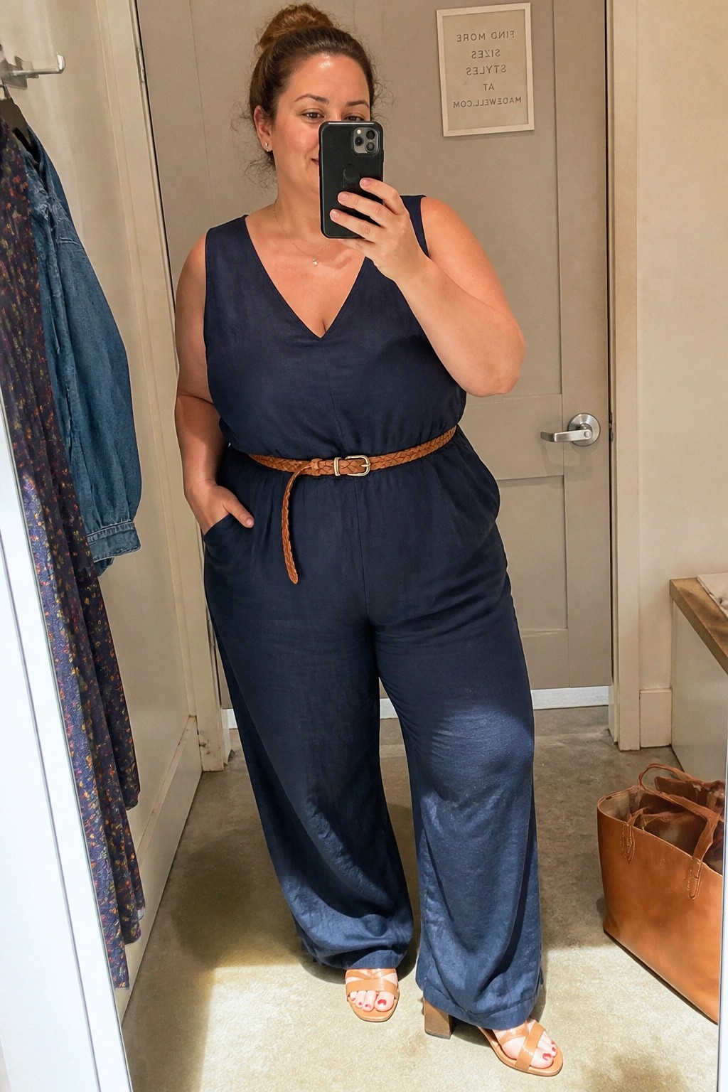 Woman in navy jumpsuit with brown belt taking mirror selfie in fitting room