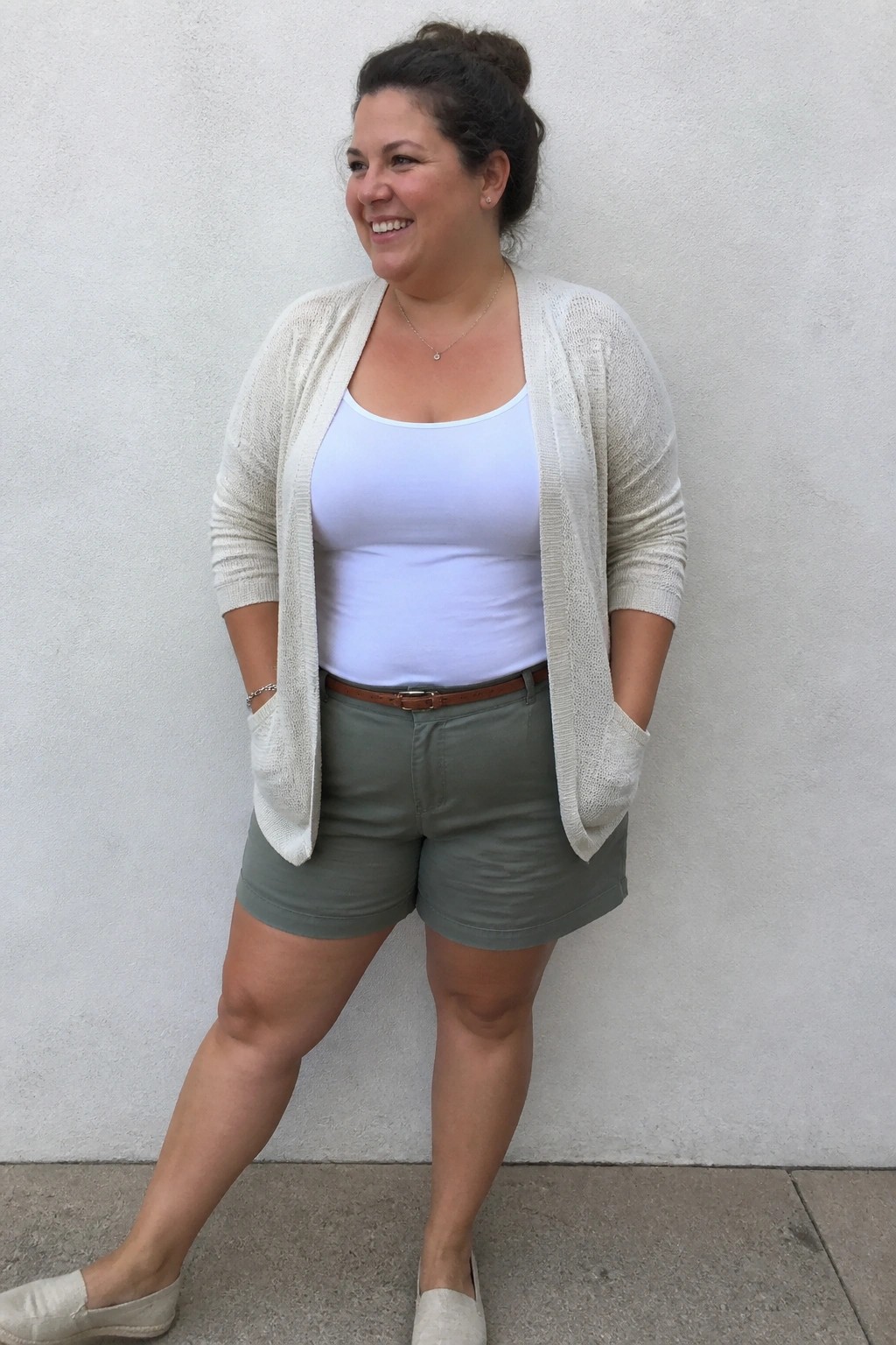 Smiling woman in open beige cardigan, white tank top, and green shorts.