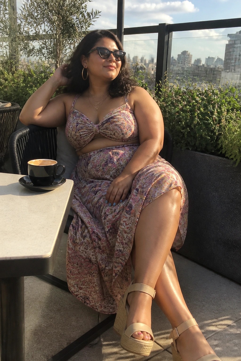 Woman in floral crop top and skirt seated on sunny balcony with coffee