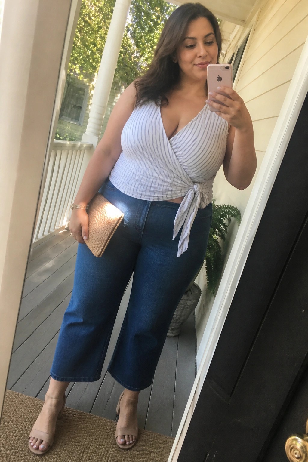 Woman in striped wrap top and wide-leg jeans taking mirror selfie on porch