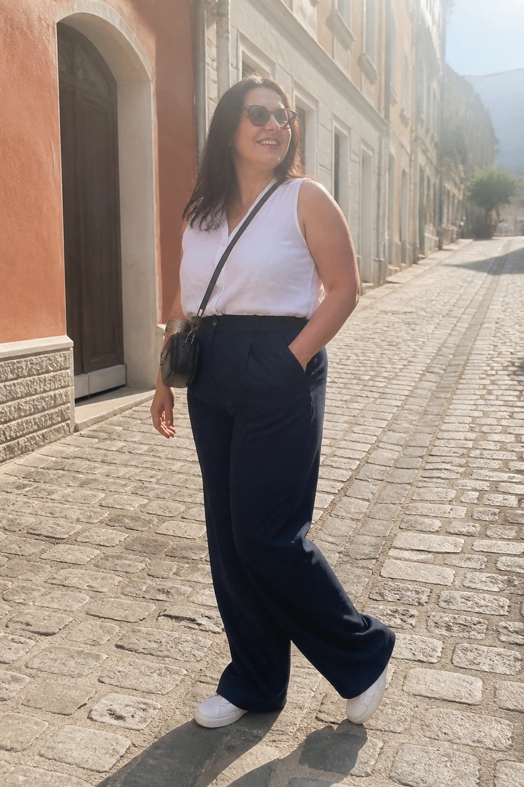 Plus size woman wearing a white sleeveless button front top tucked into navy wide leg trousers with white sneakers and a small black crossbody bag standing on a cobblestone street.