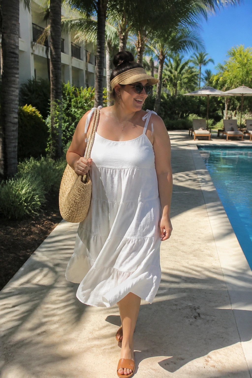Woman in white tiered dress and visor walking poolside with straw bag