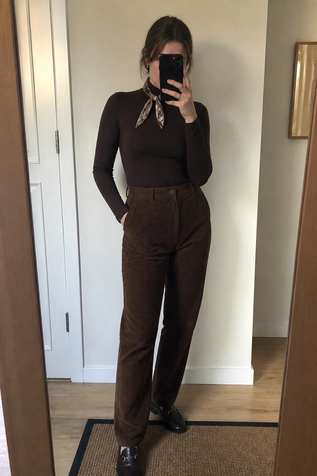 Woman wearing brown corduroy trousers with a matching ribbed long sleeve top, patterned scarf tied at the neck, and black leather loafers while standing in front of a mirror.