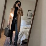 Woman taking mirror selfie in long camel wool coat over black turtleneck sweater black straight jeans black leather belt and black ankle boots while holding large black tote bag