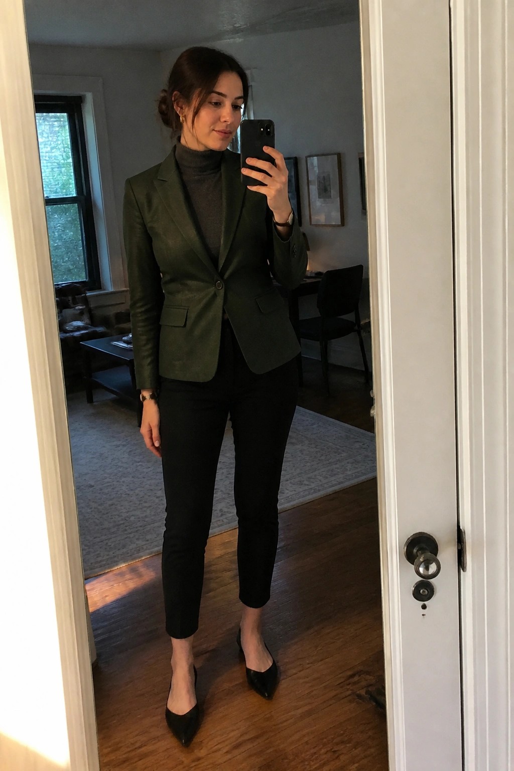 Woman in a dark green leather blazer over a gray turtleneck paired with black slim ankle pants and pointed black heels, holding a phone for a mirror selfie.
