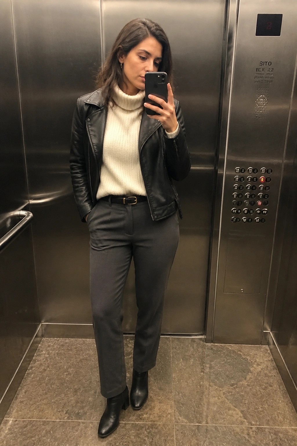 Woman in an elevator mirror selfie wearing a black leather moto jacket over a cream cable-knit turtleneck sweater, paired with grey straight-leg trousers, a black belt, and black pointed-toe ankle boots.
