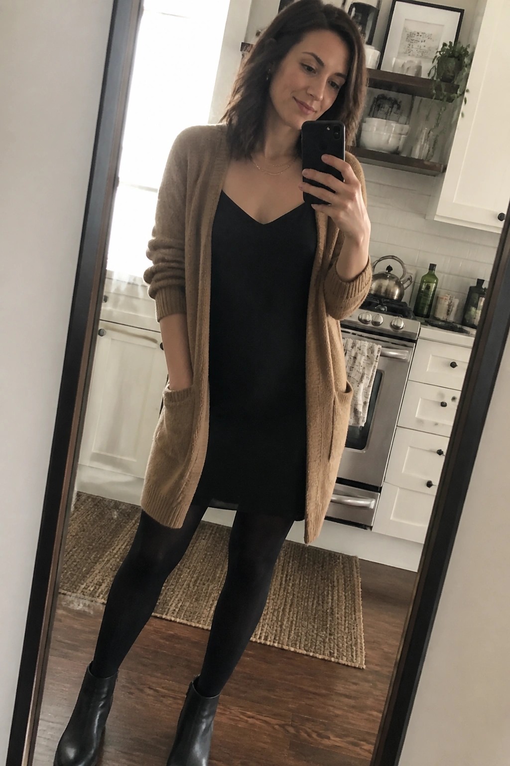 Woman in tan cardigan over black dress taking mirror selfie in kitchen