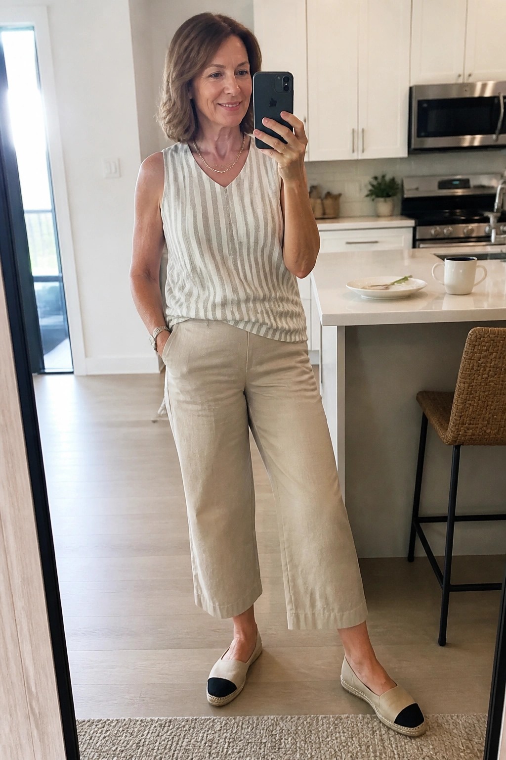 Woman over 50 in a mirror selfie wearing cropped wide-leg beige linen pants, a sleeveless white and beige striped top, layered necklaces, and beige espadrille flats with black toe caps.