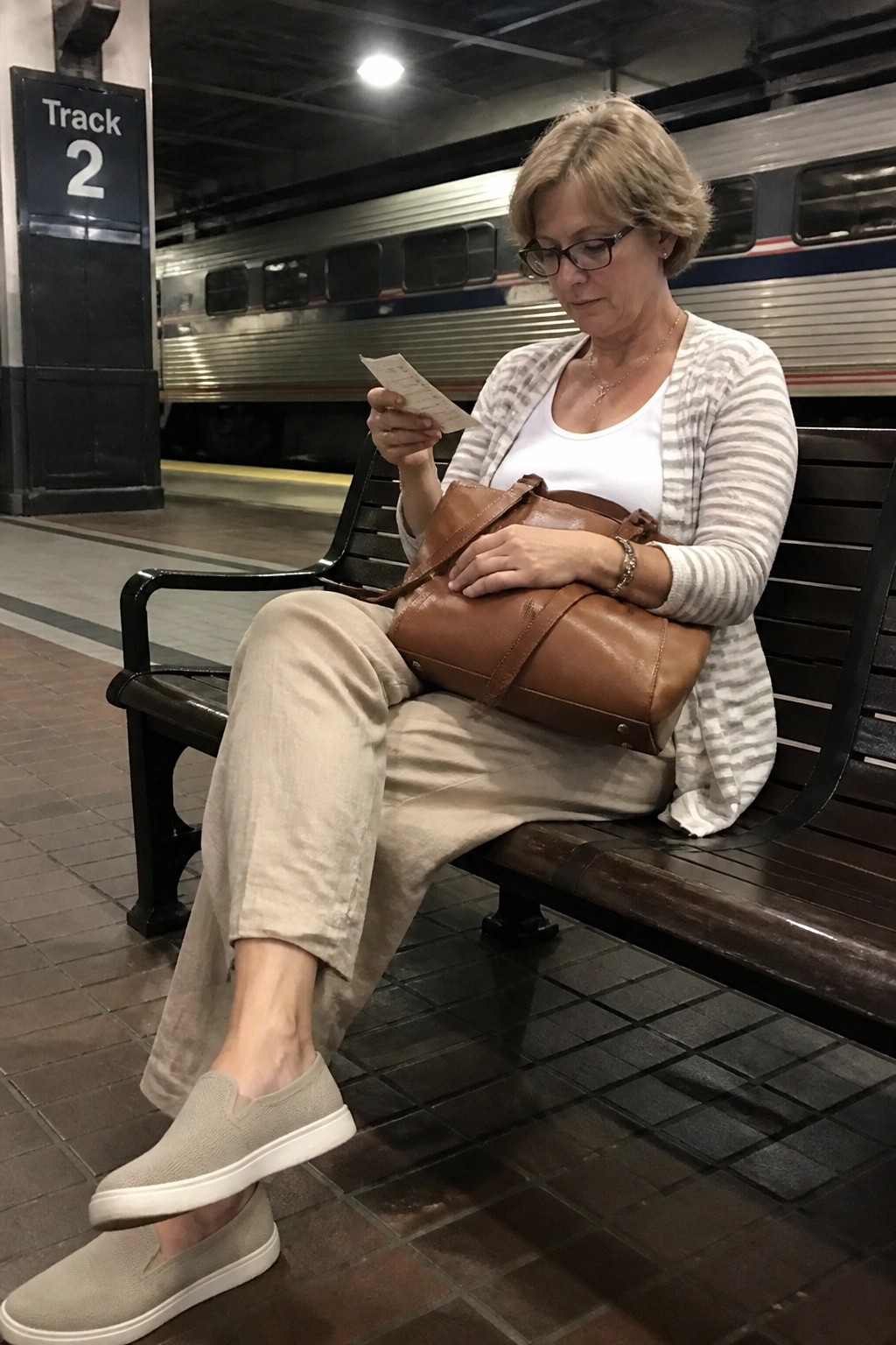 Woman in striped cardigan reading paper on bench with brown bag at train station