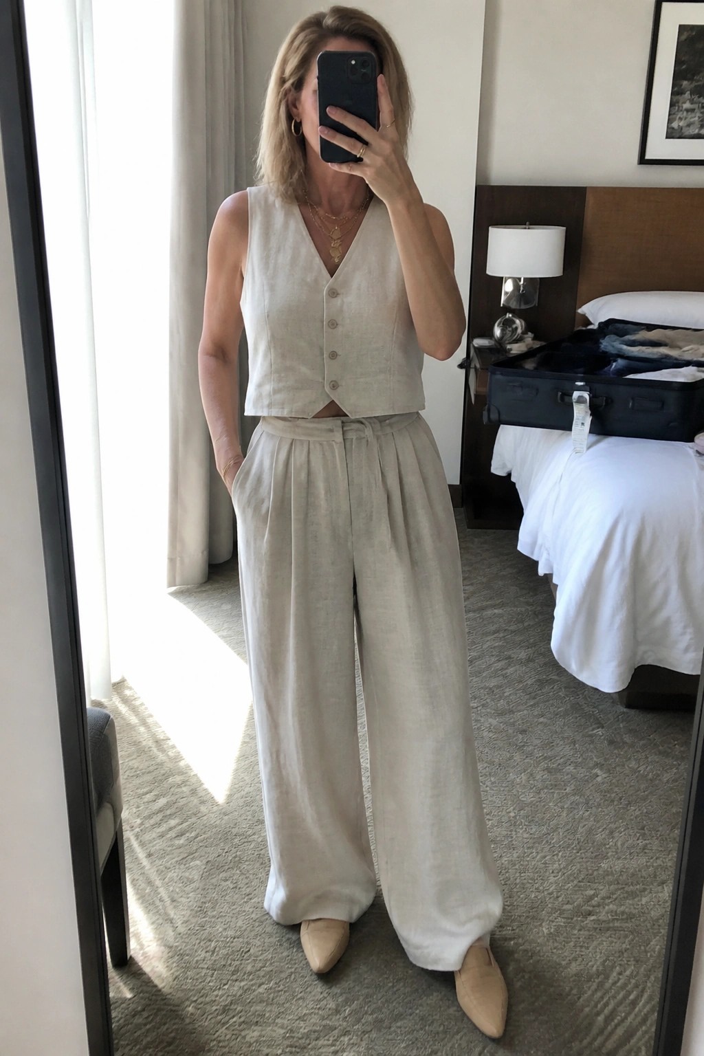 Woman in beige linen vest and wide-leg pants taking mirror selfie in hotel room.