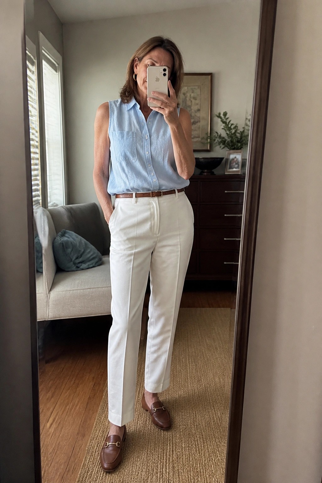 Woman over 50 wearing a light blue sleeveless button up shirt tucked into white pants with a brown belt and brown loafers while taking a mirror selfie.