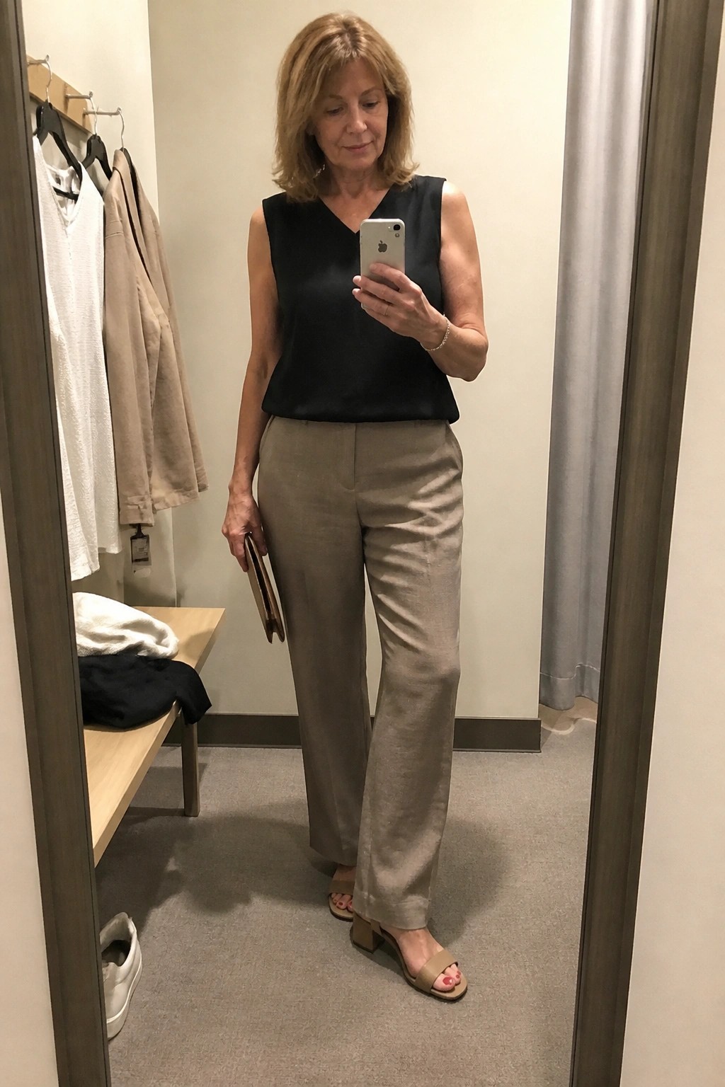 Woman over 50 wearing a black sleeveless V-neck top tucked into wide-leg beige linen pants with matching block-heel sandals while holding a phone and small clutch in a fitting room mirror selfie.