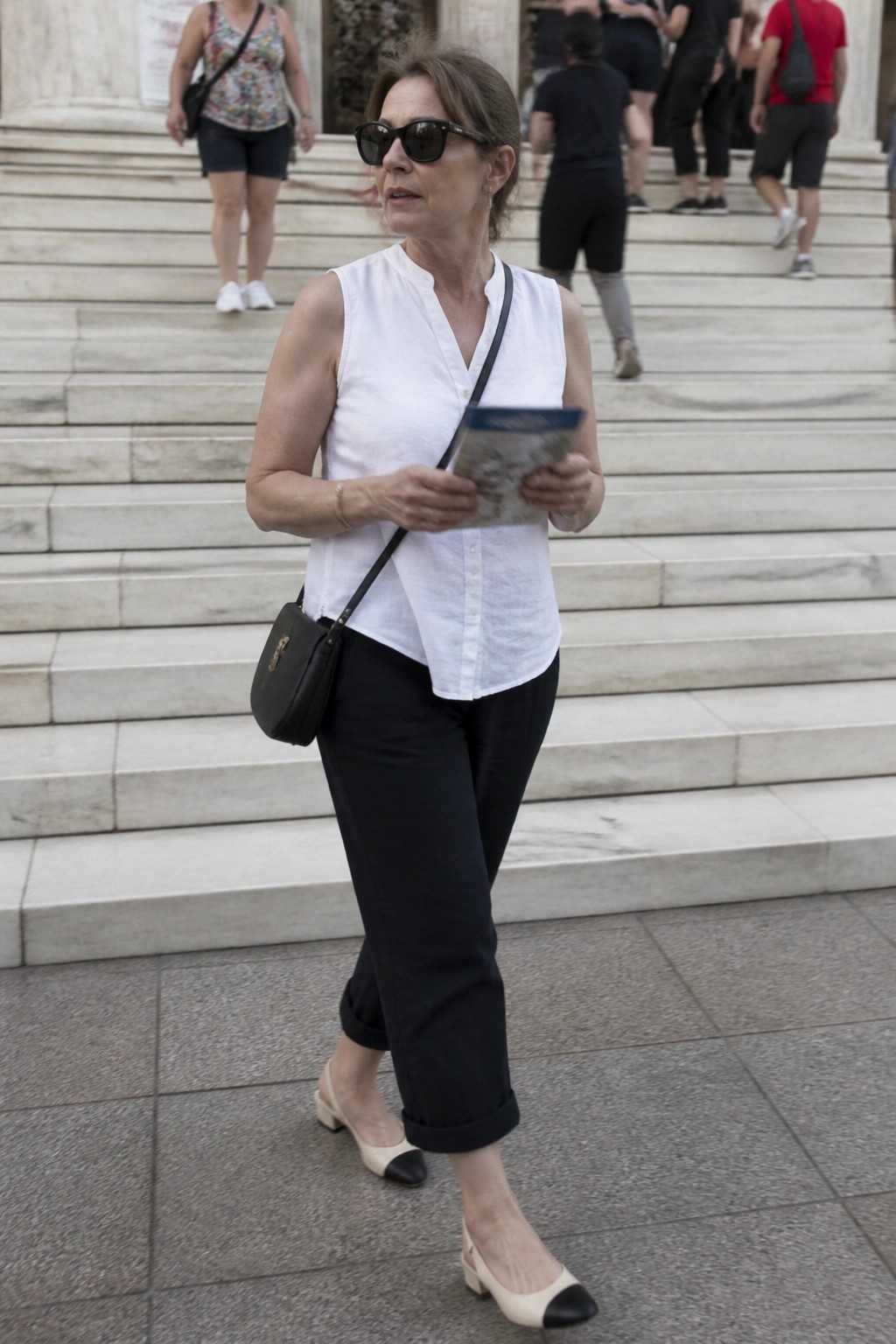 Woman over 50 in white sleeveless button front linen top tucked into black cropped cuffed pants with beige and black cap toe flats plus black crossbody bag while holding a small booklet on stone steps.
