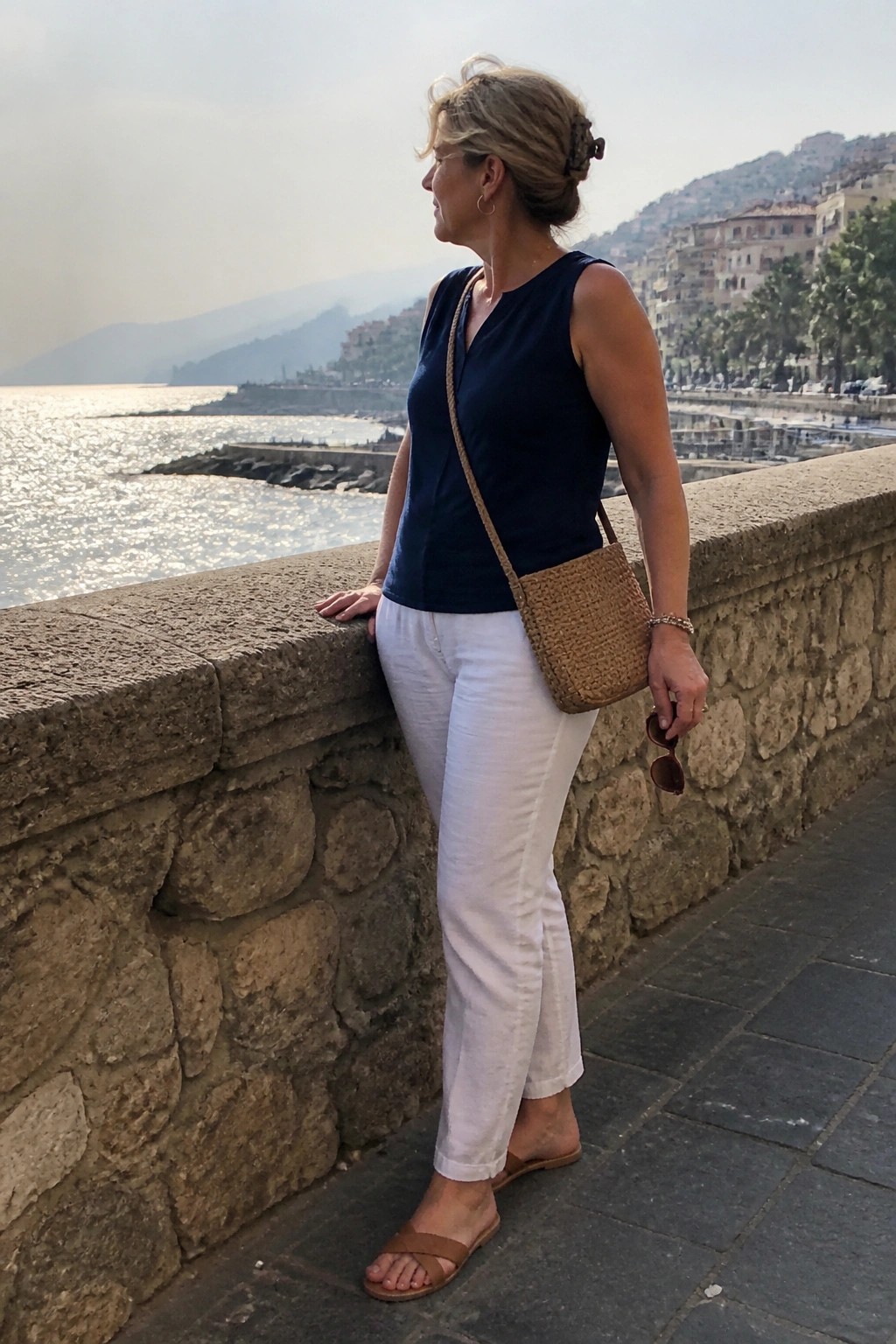 Woman wearing white straight-leg pants with a navy sleeveless V-neck top, brown woven crossbody bag, and matching flat sandals, holding sunglasses in one hand.