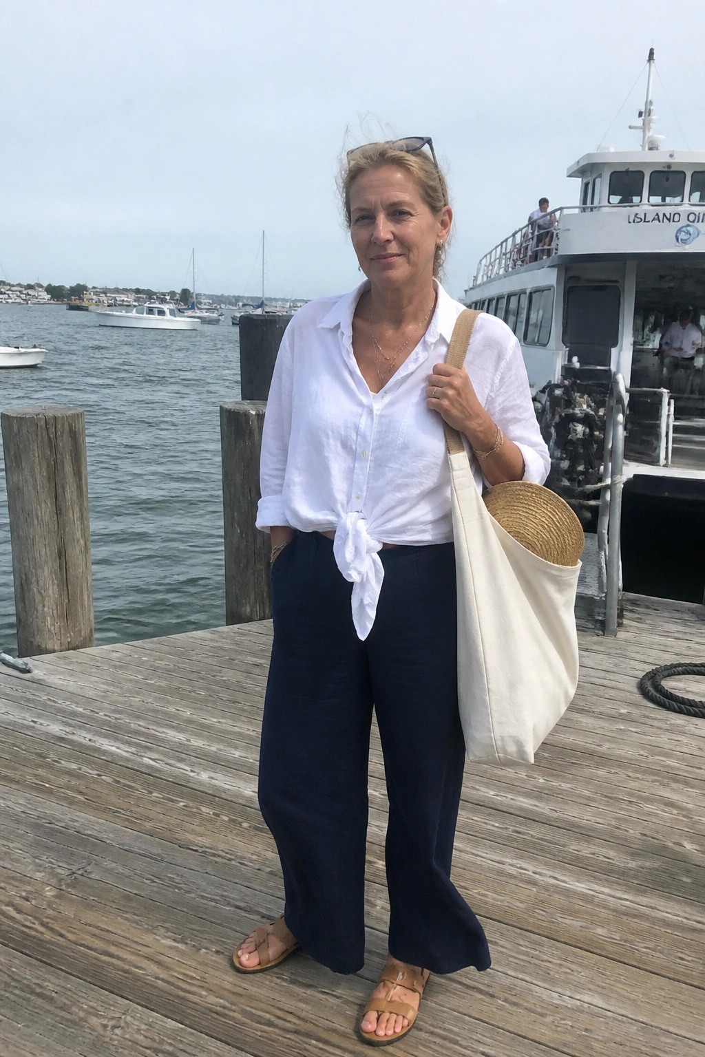 Woman over 50 wearing a white linen button up shirt knotted at the waist with navy wide leg linen pants brown strappy sandals and layered gold necklaces while carrying a canvas tote bag containing a straw hat.