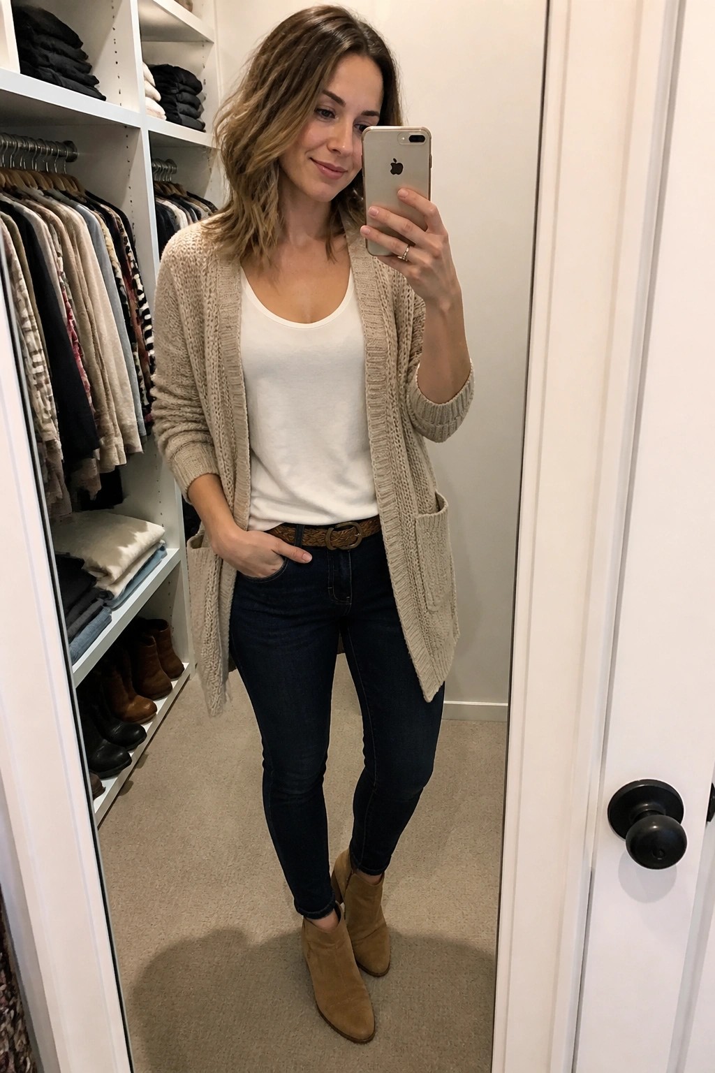Woman in beige cardigan and jeans taking iPhone mirror selfie in walk-in closet