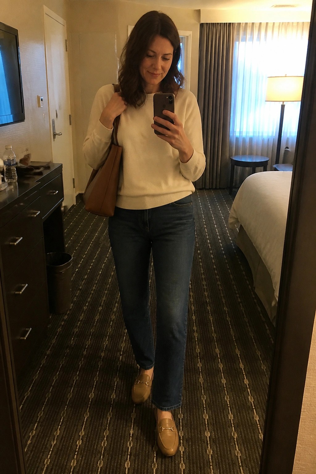 Woman wearing a cream sweater tucked into blue straight-leg jeans with beige horsebit loafers and carrying a brown leather tote bag while taking a mirror selfie.