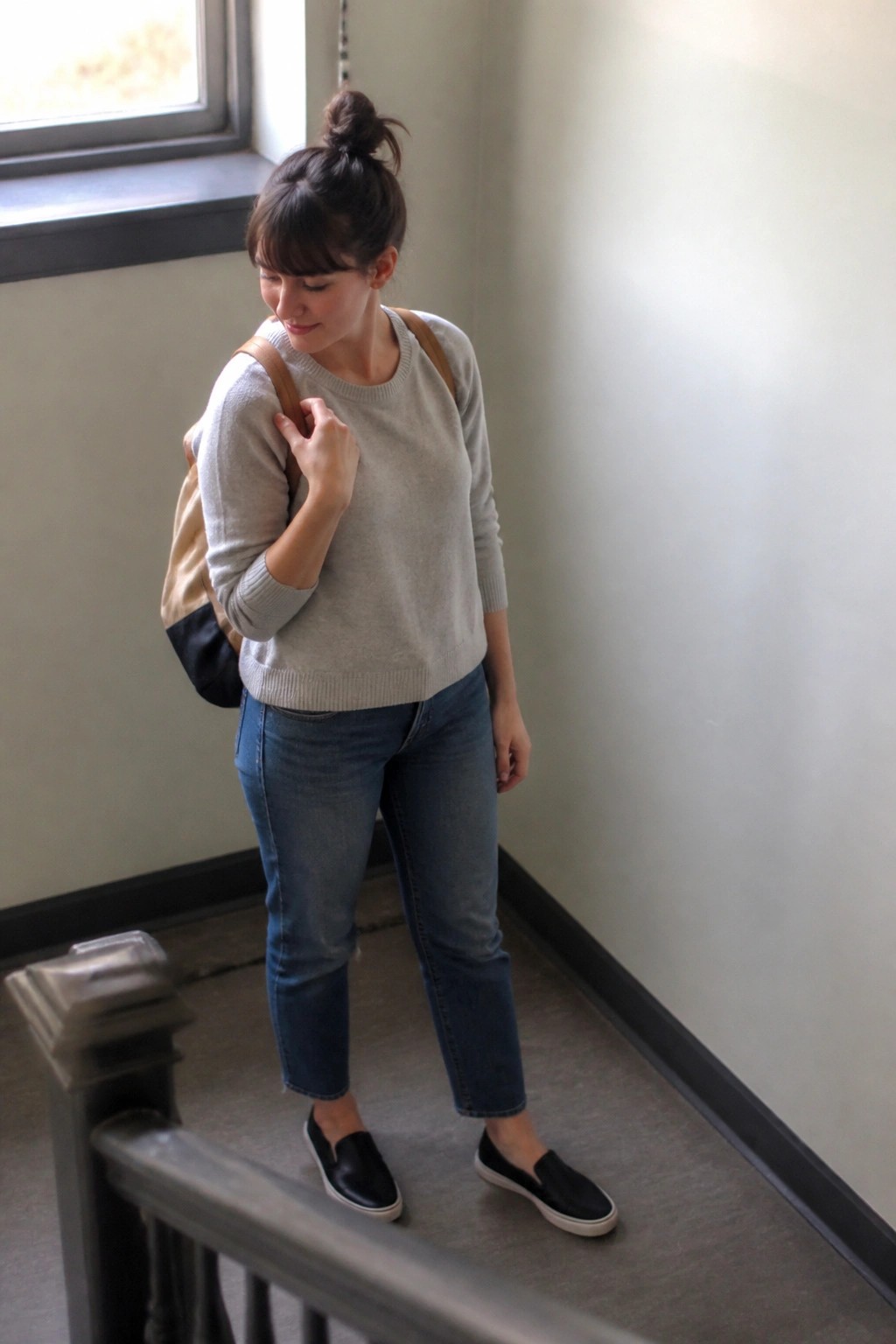 Woman in gray sweater and jeans carrying backpack on indoor stairs
