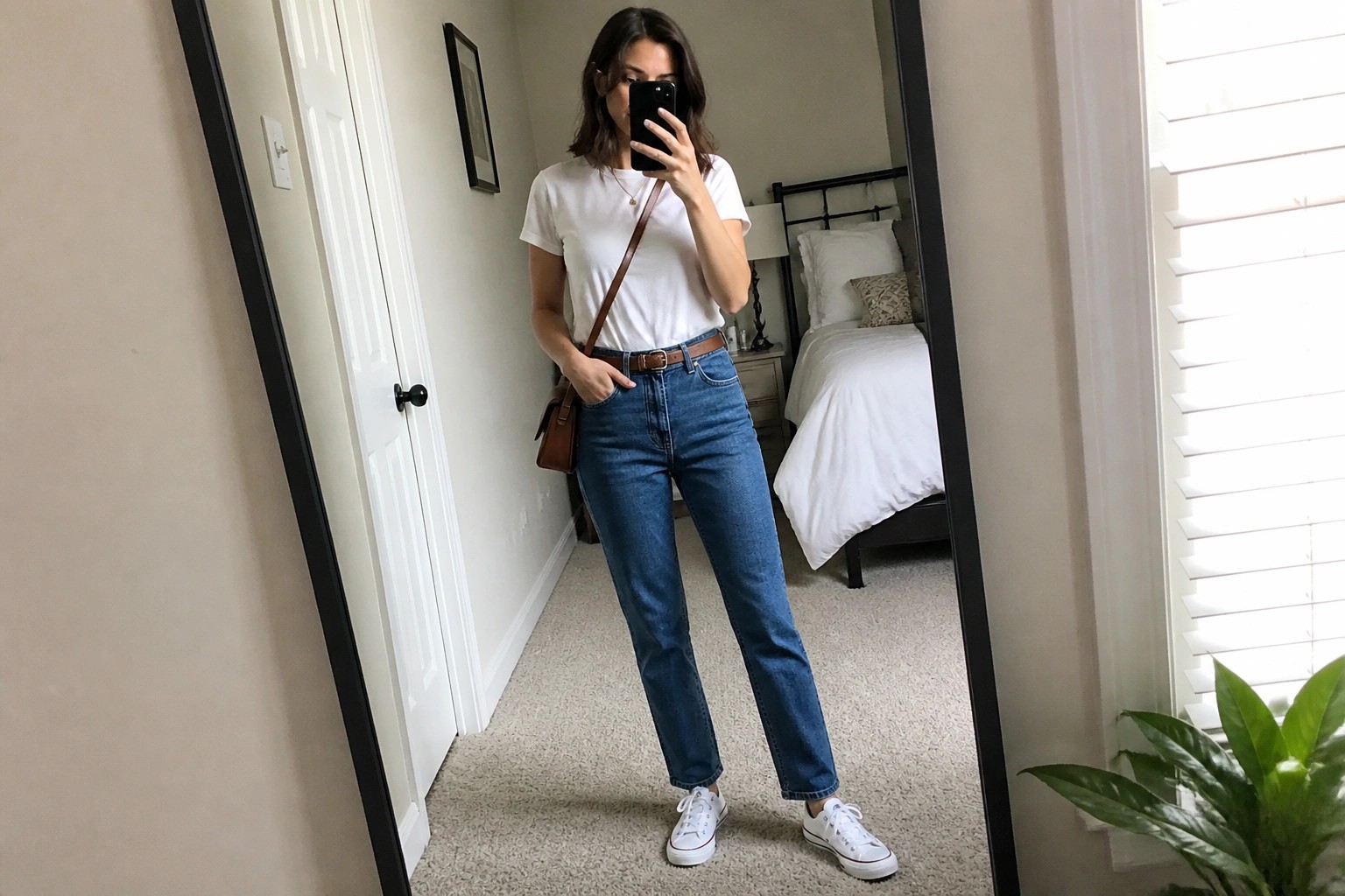 Woman wearing a white short sleeve t-shirt tucked into blue straight leg jeans with a brown leather belt, white sneakers, a brown crossbody bag, and a thin gold necklace.
