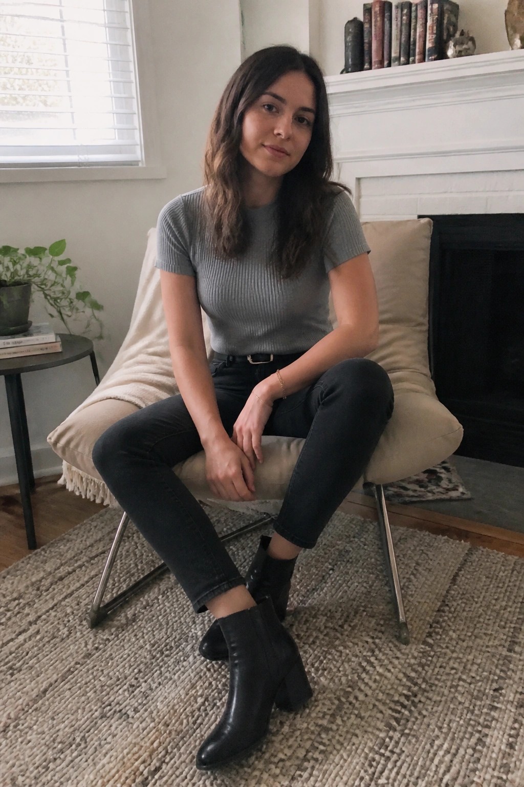Woman in gray top and black boots seated in beige armchair by fireplace