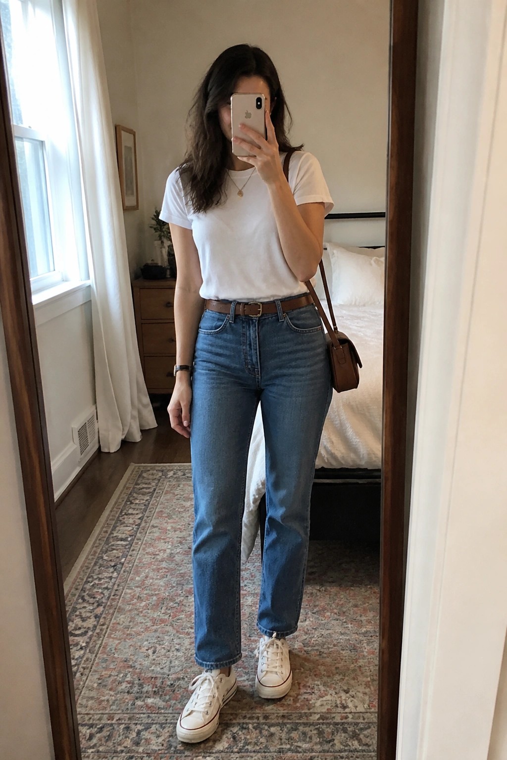 Woman wearing a white short sleeve t-shirt tucked into blue straight leg jeans with a brown leather belt, white sneakers, a brown crossbody bag, and a thin gold necklace.