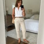 Woman over 50 in a white sleeveless linen shirt tucked into beige trousers with a brown belt and pointed flats while holding a phone and woven straw bag.