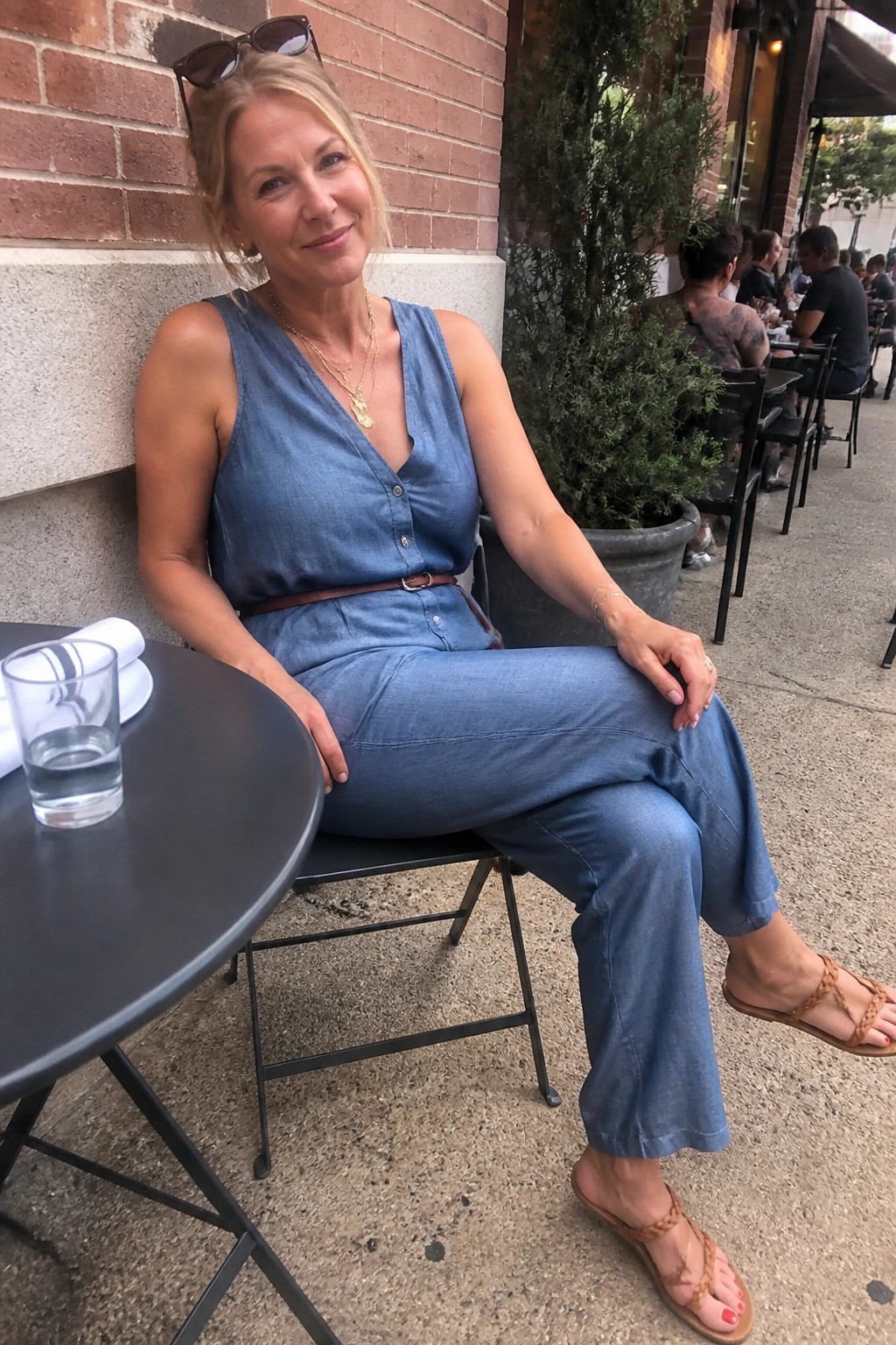 Blonde woman in blue denim jumpsuit seated at outdoor cafe table with water glass